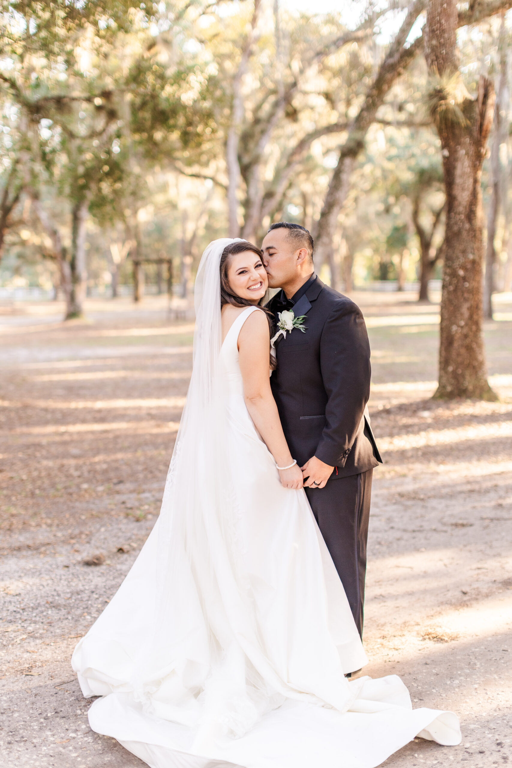 An enchanting autumn wedding scene at Chandler Oaks Barn in St. Augustine, Florida. A radiant couple stands beneath the sprawling branches of majestic oak trees near a charming white gazebo adorned with seasonal florals. The bride wears a flowing white gown, holding a bouquet of vibrant fall blooms, while the groom is dressed in a sharp suit. Warm, golden sunlight filters through the trees, illuminating the serene setting and highlighting the romantic, classic charm of the sunny Florida afternoon.